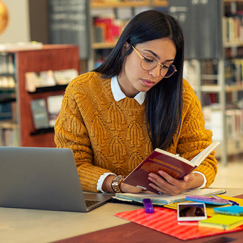 Young lady in a library reading from a notebook while sitting in front of a laptop computer.