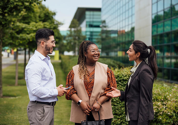 Three diverse colleagues smiling and talking in an outdoor setting.