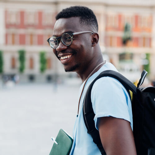 Young adult with eyeglasses walking on street with a backpack and notebooks in his hand 