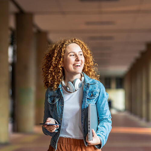 Student wearing a jean jacket, with headphones around her neck, a cell phone in one hand, and a laptop under her opposite arm.