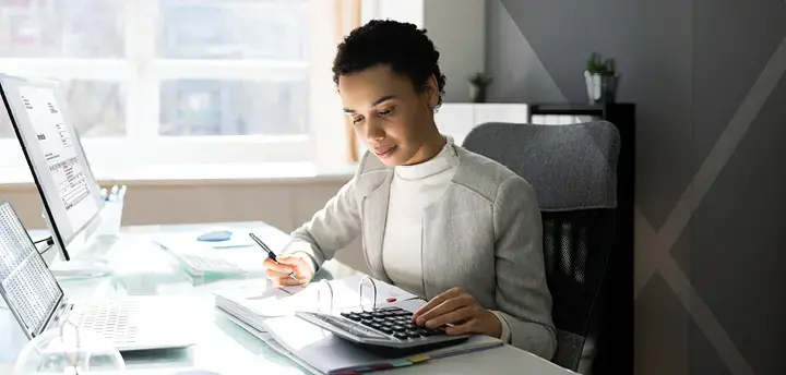 Young professional sitting at a desk and looking down at a calculator with pen in hand.