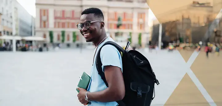 Young adult with eyeglasses walking on street with a backpack and notebooks in his hand 