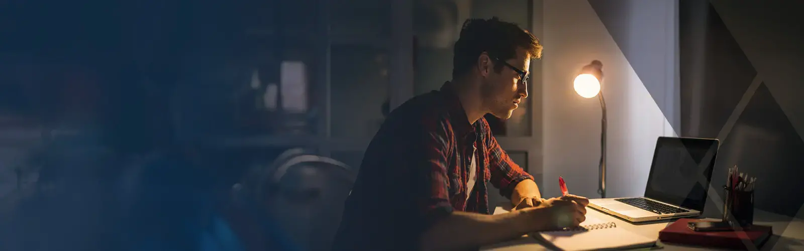 College student wearing eyeglasses, sitting at desk studying, and writing down notes using laptop 