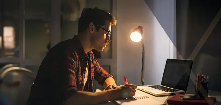 College student wearing eyeglasses, sitting at desk studying, and writing down notes using laptop 