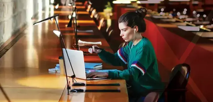 Student sitting at a library table, looking at a laptop 