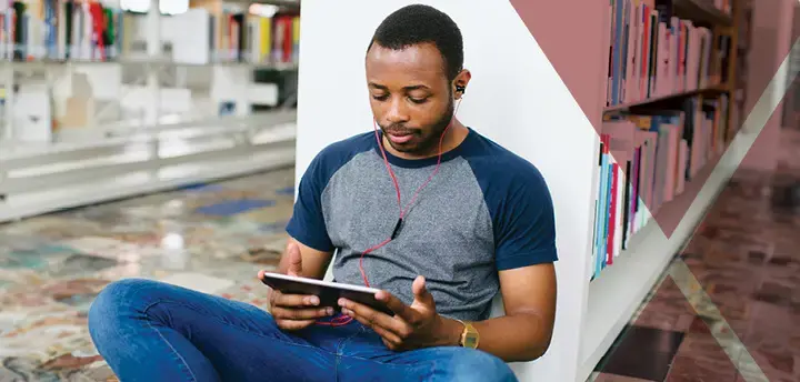 A student sits in library on tablet with earphones in 