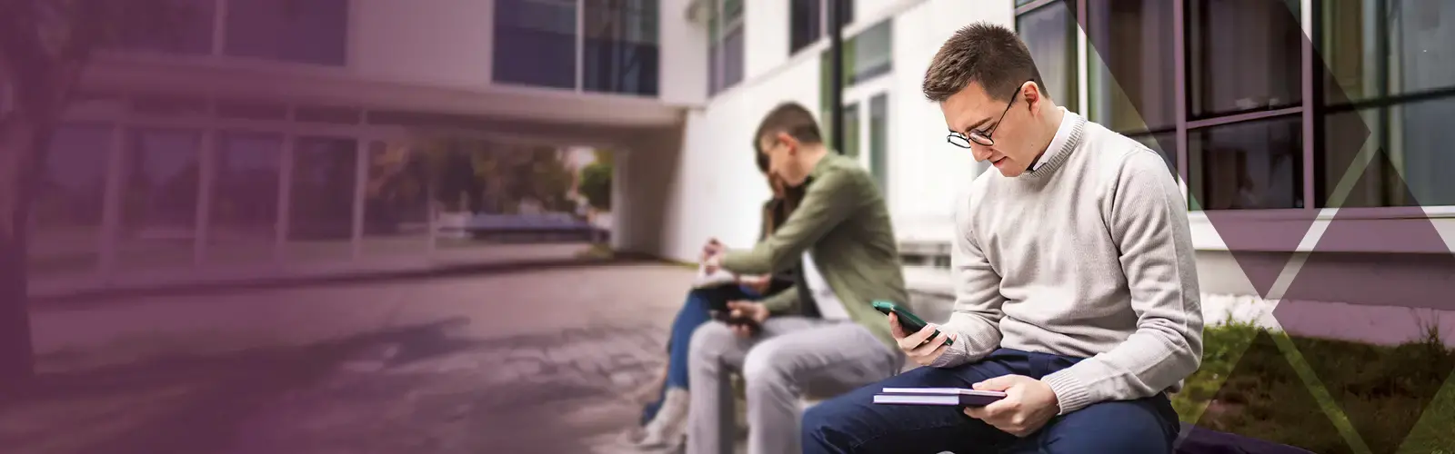College students sitting on a wall outside a building; one is looking at his phone
