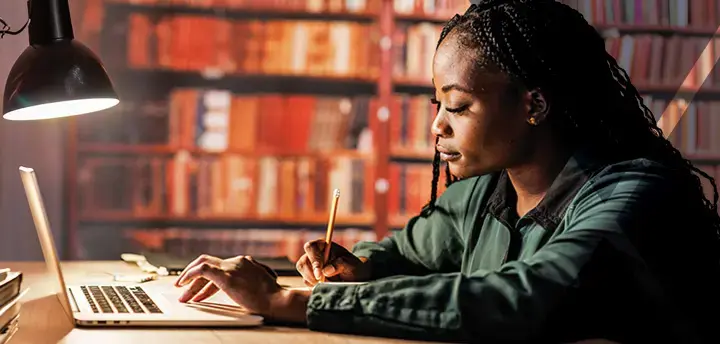 Diligent young female student sitting at table at the library, using laptop and writing notes while studying for Washington Bar Exam.